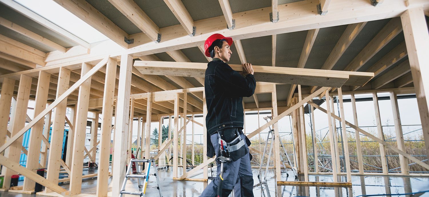 En byggmontør med rød vernehjelm på hode bærer to treplanker på skulderen i en byggeplass. Foto.